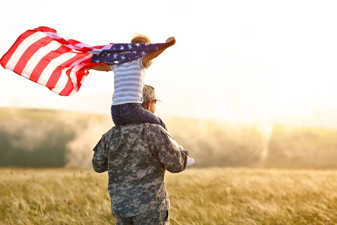A veteran in uniform carrying a child with an American flag through a golden field