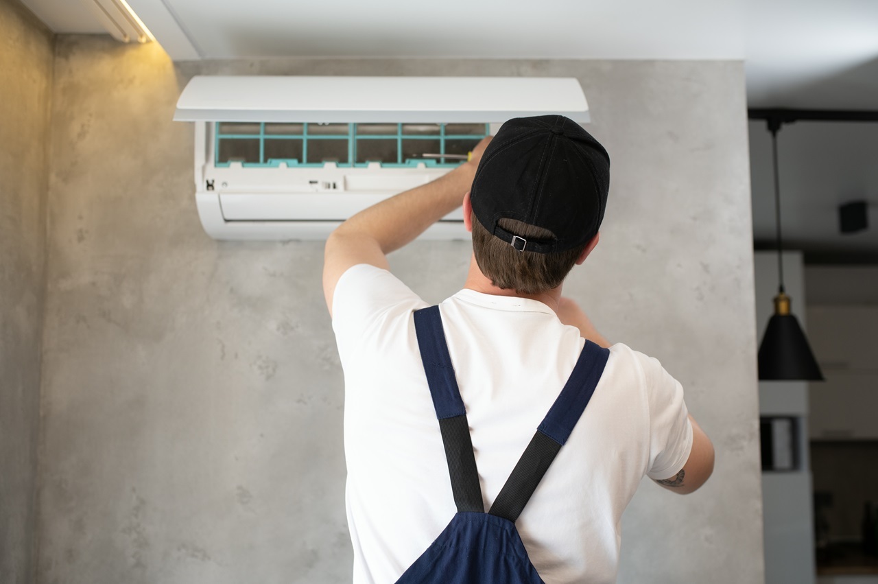 A man in overalls and a hat repairs an air conditioner.