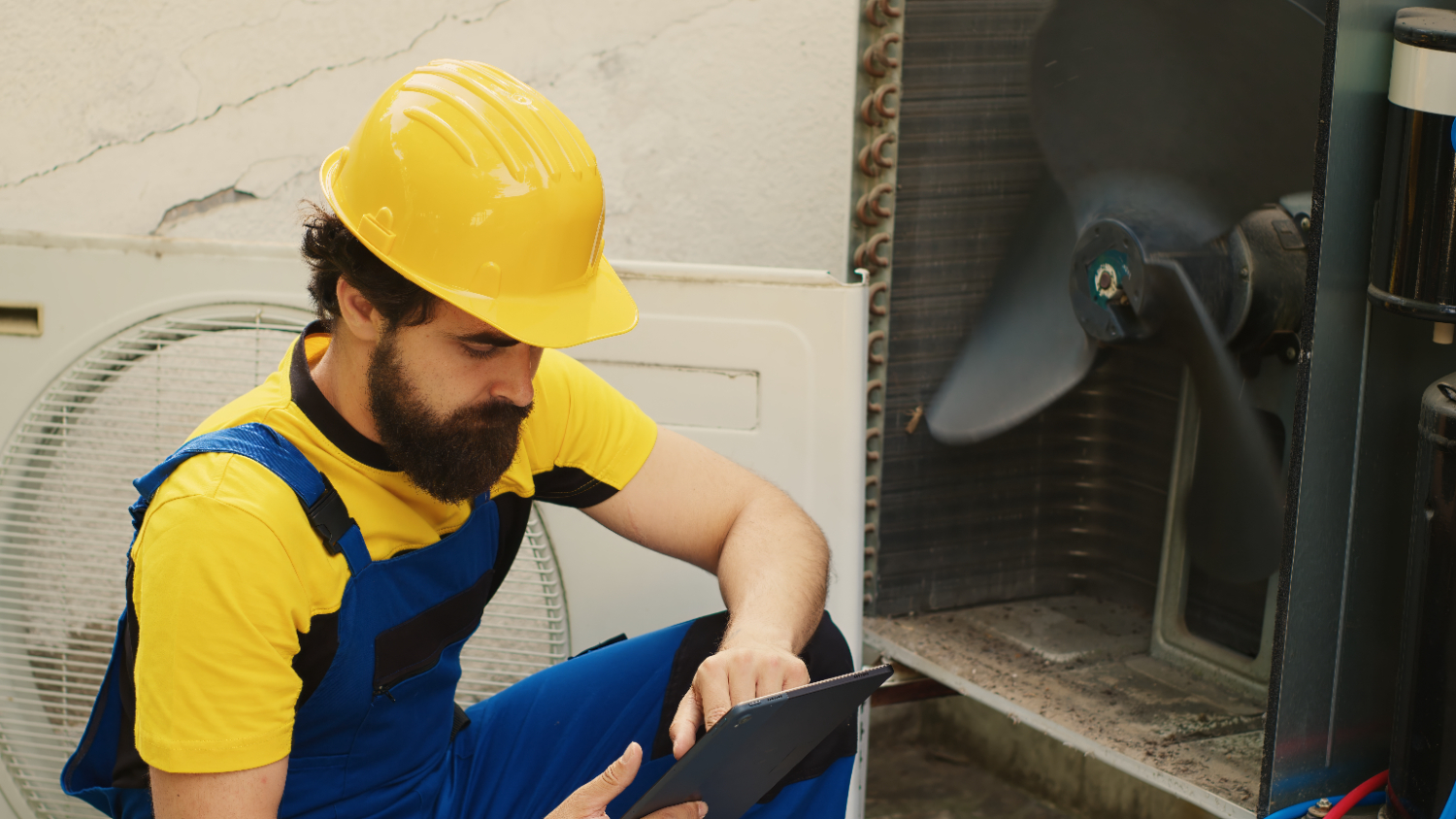 A man in overalls and a hard hat holds a tablet computer, focused on his work.